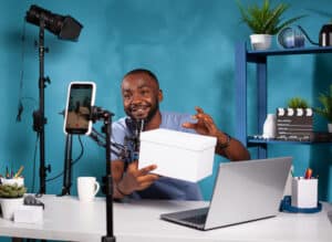 influencer-presenting-white-mockup-gift-fans-looking-live-video-podcast-setup-vlogging-studio-smiling-vlogger-holding-product-box-hosting-online-giveaway-front-recording-smartphone-300x219 Your Ultimate Guide On Podcast How To Start: From Concept To Launch